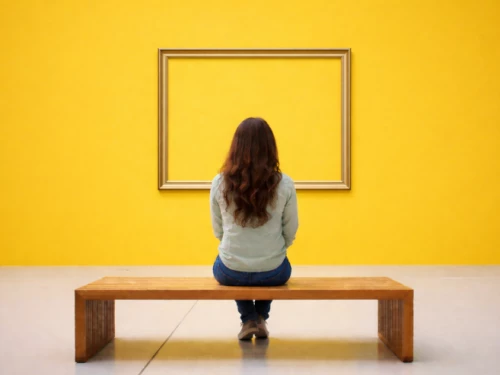 femme assise sur un banc devant un mur jaune. Face à elle un cadre nu.